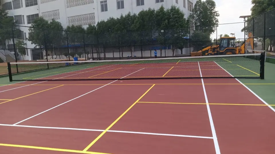 Outdoor court in red with a green surround, white tennis lines, additional yellow line markings and a tennis net, with a building and an excavator behind.
