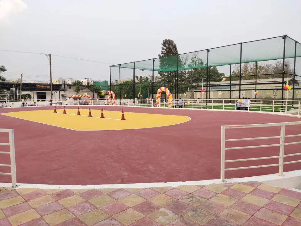 Outdoor skating rink with a maroon track around a yellow centre, cones set out and steel railings, with event decorations and a netted box cricket arena behind.