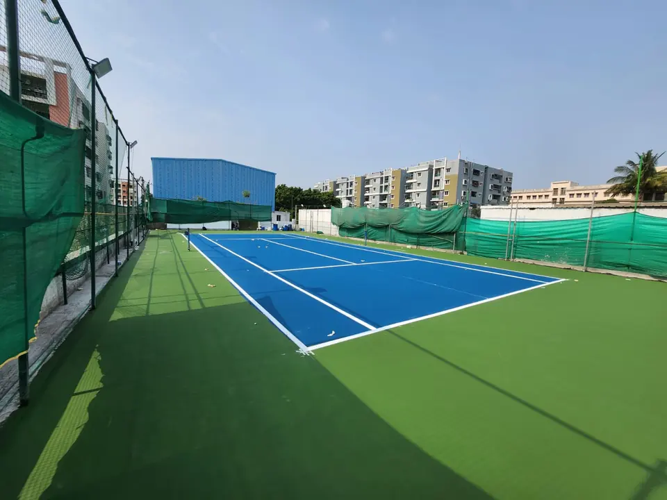 Outdoor tennis court with a blue playing area and wide green surround, edged by green shade netting and floodlight poles, with apartment blocks behind.