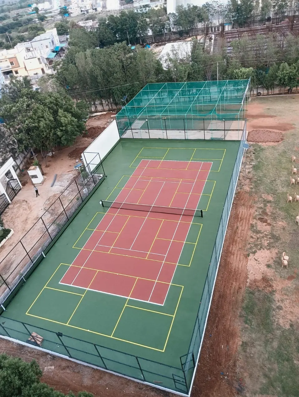 Aerial view of a fenced court with a red playing area marked for tennis, yellow lines for other games on the green surround, and a netted practice cage beyond.