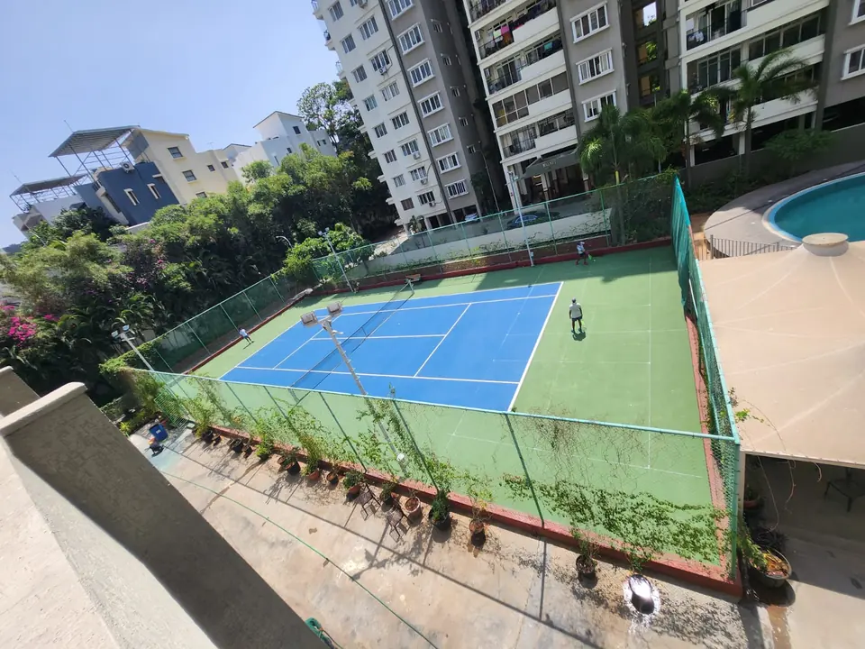 Aerial view of a fenced outdoor tennis court with a blue playing area and green surround, beside a swimming pool in a residential complex, with players on court.