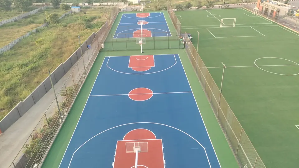 Aerial view of two full outdoor acrylic basketball courts in blue with red keys and centre circles, fenced, beside an artificial turf football pitch with goals.