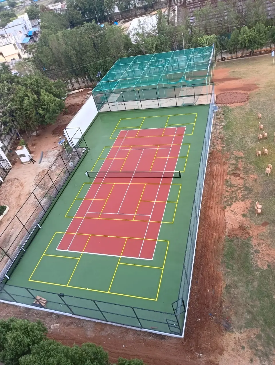 Aerial view of a fenced outdoor court with a red pickleball playing area, yellow lines for other sports and a netted practice cage alongside.