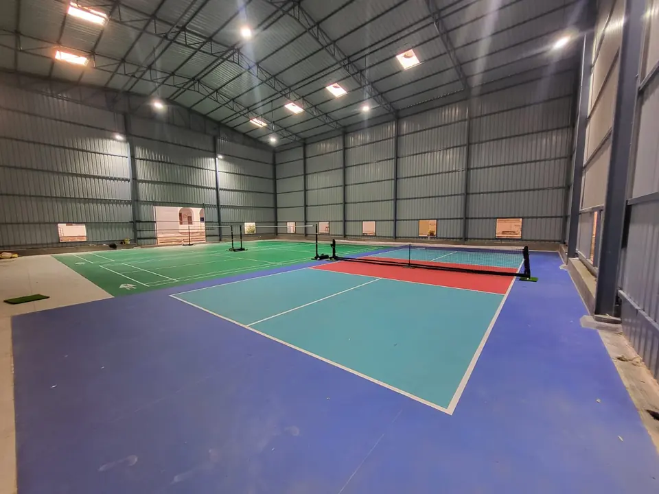 Indoor pickleball court with a net inside a tall steel shed with high-bay lights, next to an indoor badminton court.