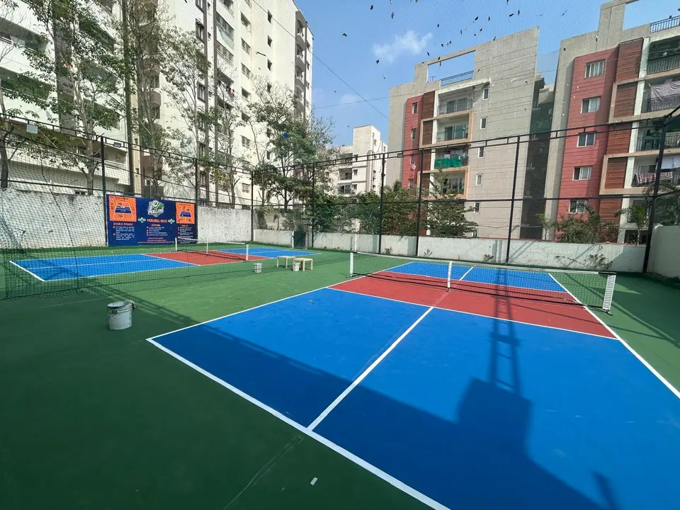 Multi-court outdoor pickleball facility built by Kelysports, with blue courts, red kitchen zones and nets, overlooked by apartment buildings.