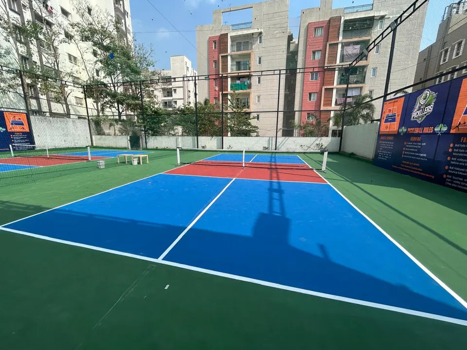 Outdoor pickleball court construction by Kelysports: blue courts with red kitchen zones and nets inside a netted enclosure beside apartment blocks.