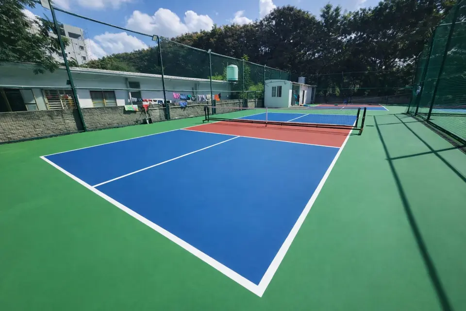 Outdoor pickleball court with blue playing areas and a red kitchen zone, enclosed by chain-link fencing among trees.