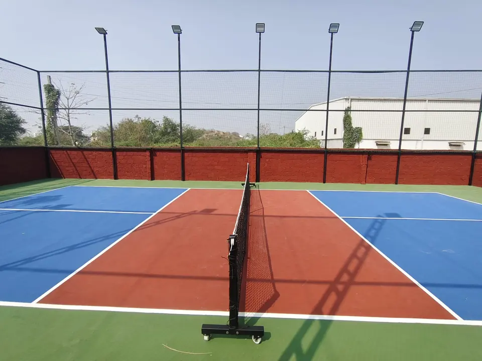Outdoor pickleball court with a red kitchen zone and net, a brick boundary wall and a row of floodlight poles.