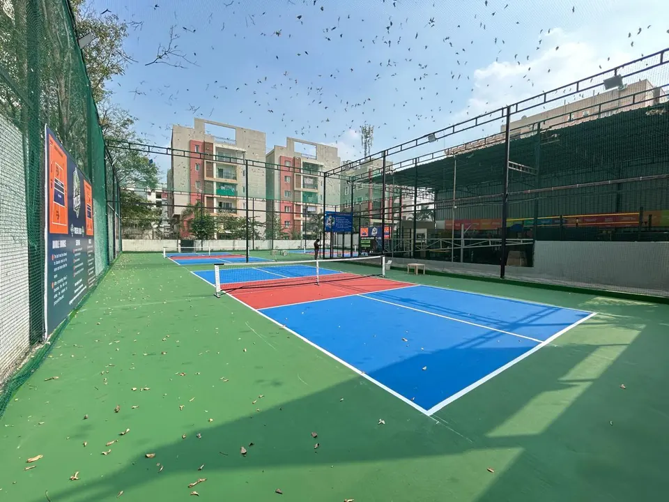 Outdoor pickleball courts inside a tall netted steel enclosure, with rule boards on the fence and apartment blocks behind.