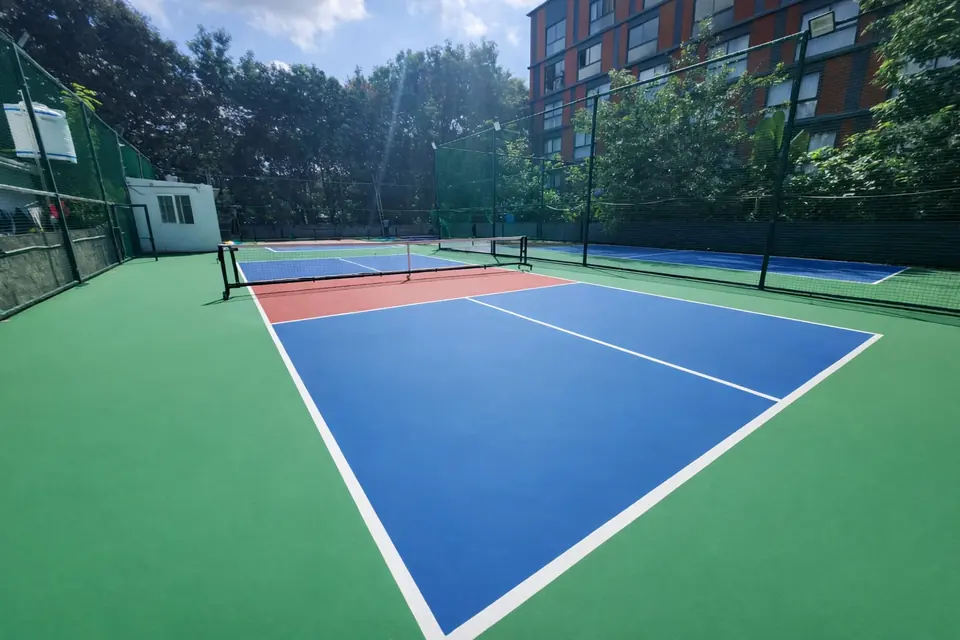 Baseline view of a fenced outdoor pickleball court, showing the blue playing area, red non-volley zone and net.