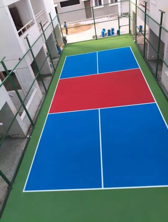 Fenced pickleball court with a blue playing area and red kitchen zone, built in a narrow space between residential buildings.
