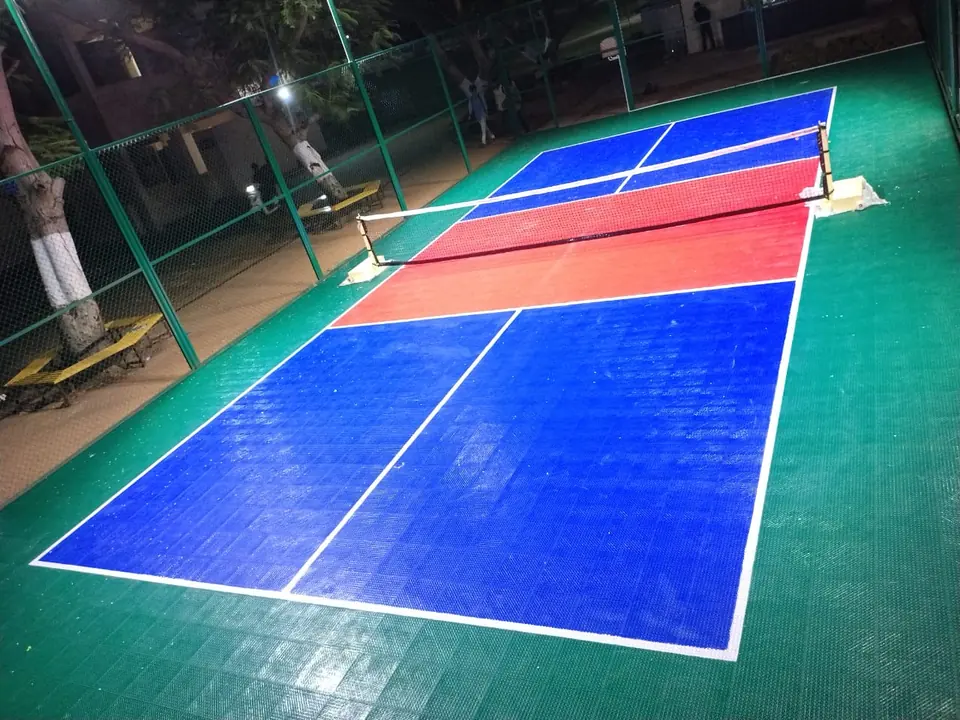 Pickleball court on blue, red and green interlocking tiles, lit for evening play, with the net up and fencing around it.