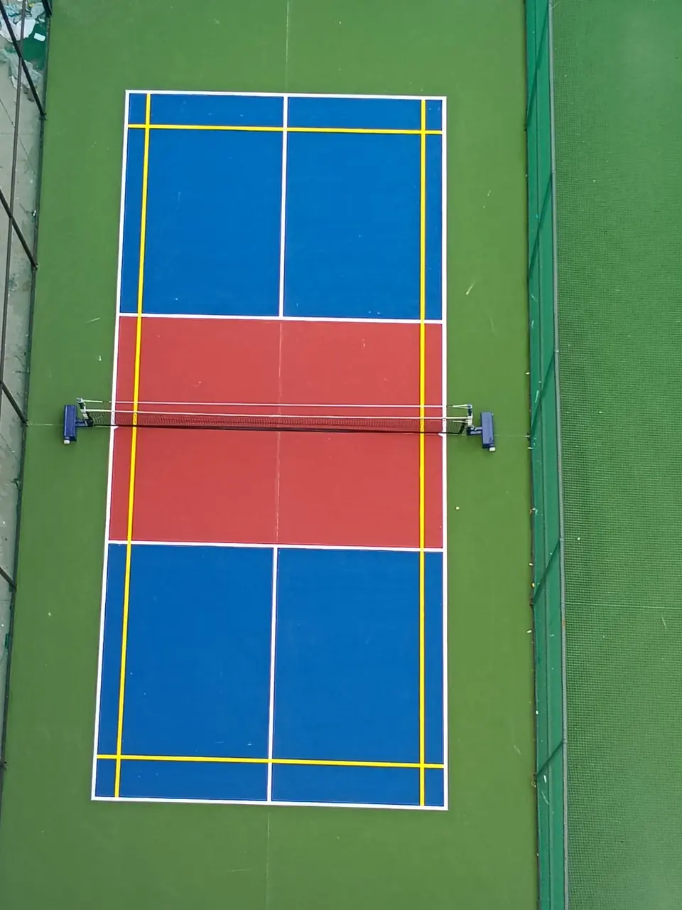 Overhead view of a pickleball court with a net and additional yellow badminton lines, set on a green surround.