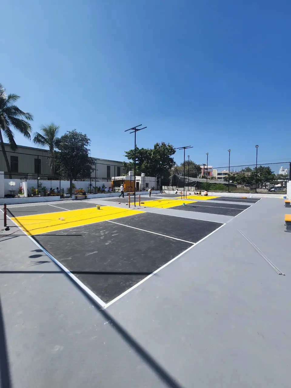 Outdoor pickleball courts being finished on a grey base, with charcoal playing areas, yellow kitchen zones and floodlight poles.