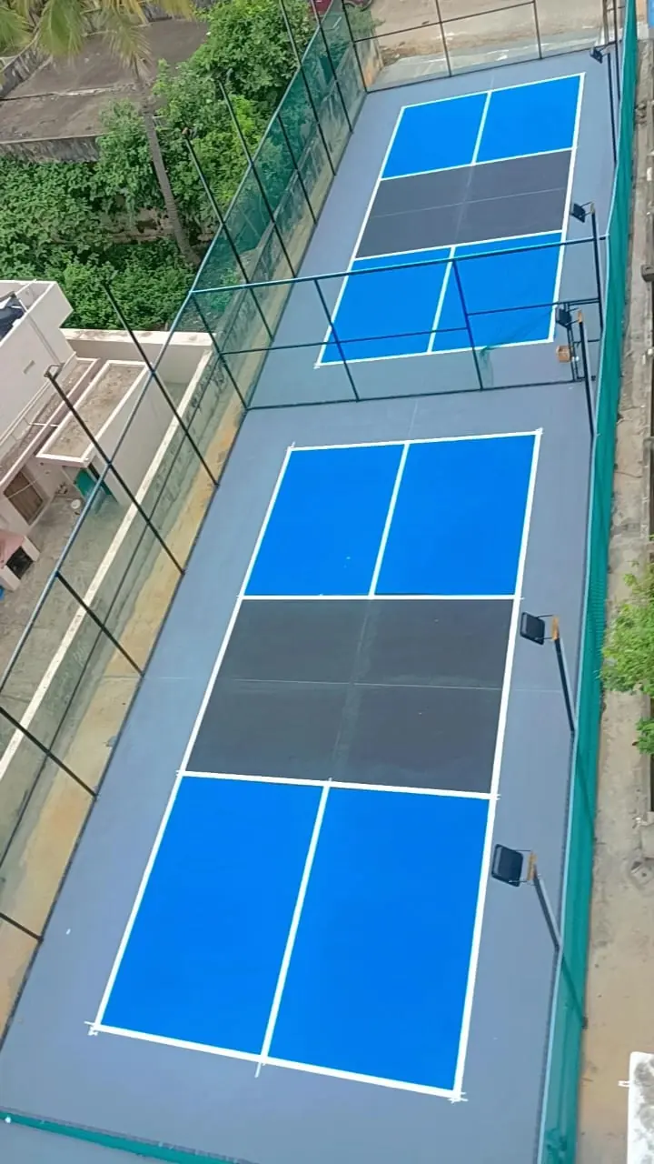 Two pickleball courts in light blue with charcoal kitchen zones on a long rooftop, fenced and fitted with floodlights.