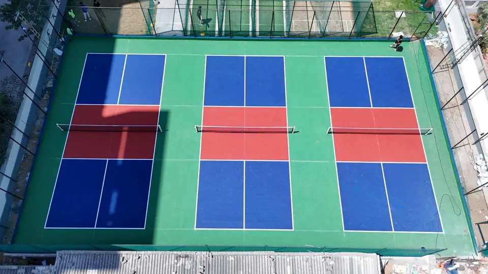 Overhead view of three pickleball courts side by side with nets, blue playing areas, red kitchen zones and a green surround inside a fenced enclosure.