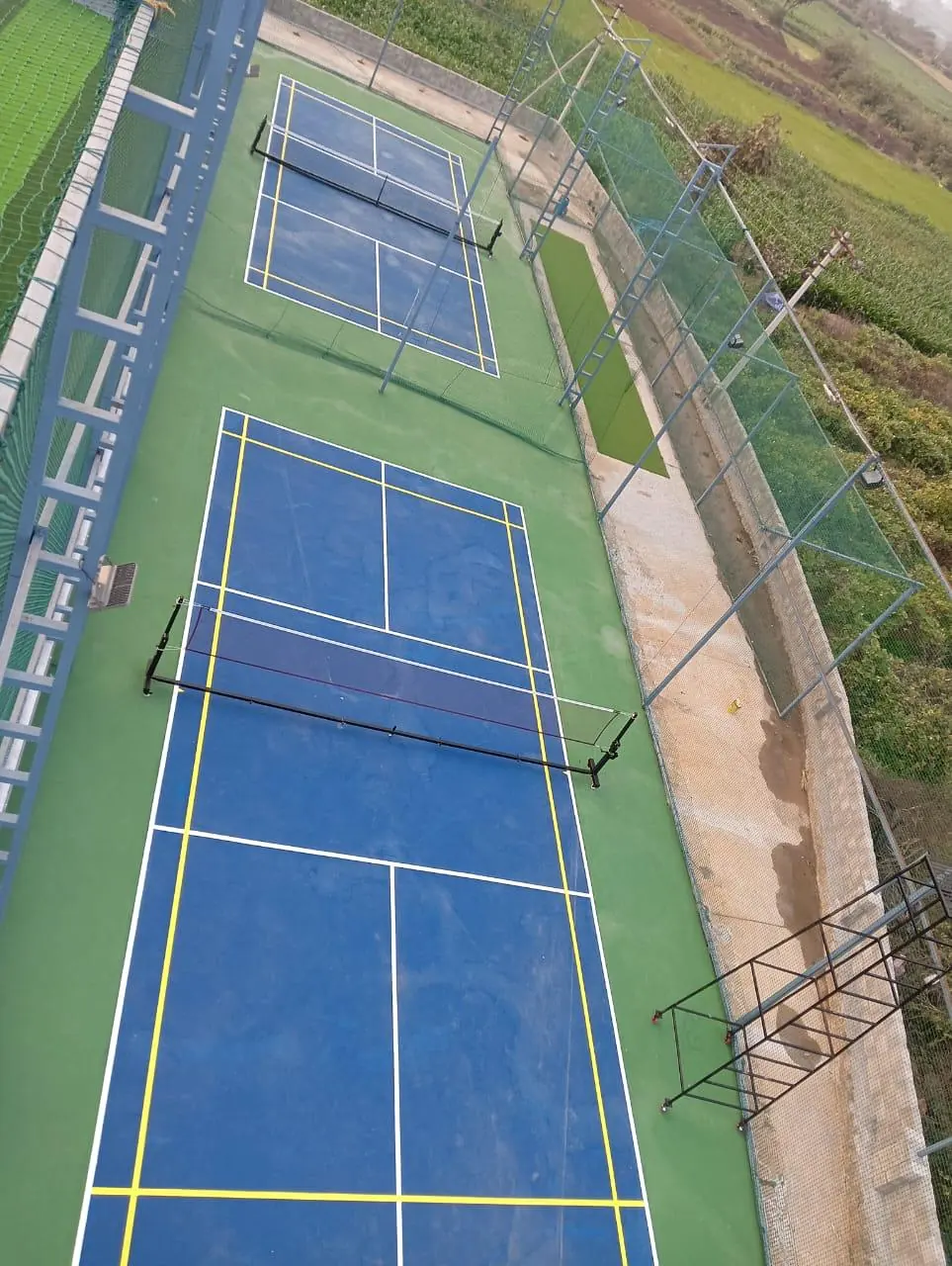 Two pickleball courts laid end to end on a long, narrow plot, with yellow badminton lines and netting along the sides.