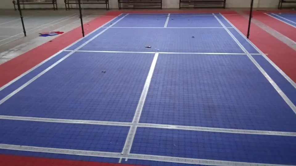 Indoor badminton court laid with blue PP interlocking tiles and a red surround, with white court lines in place and spare tiles beside the court, in an older sports hall.