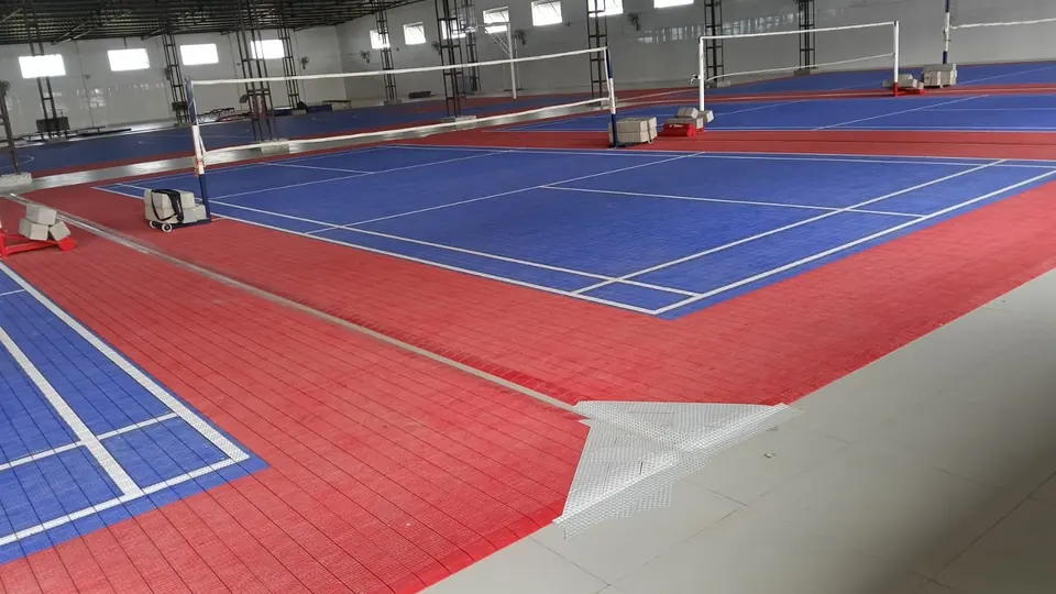 Badminton courts on blue and red PP interlocking tiles in a large multi-court hall, with net posts, an edge ramp tile and a basketball court beyond.
