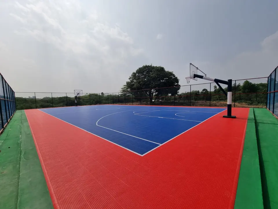 The same PP tile basketball court seen from the other end, with a pole and backboard, the red surround and green border inside chain-link fencing.