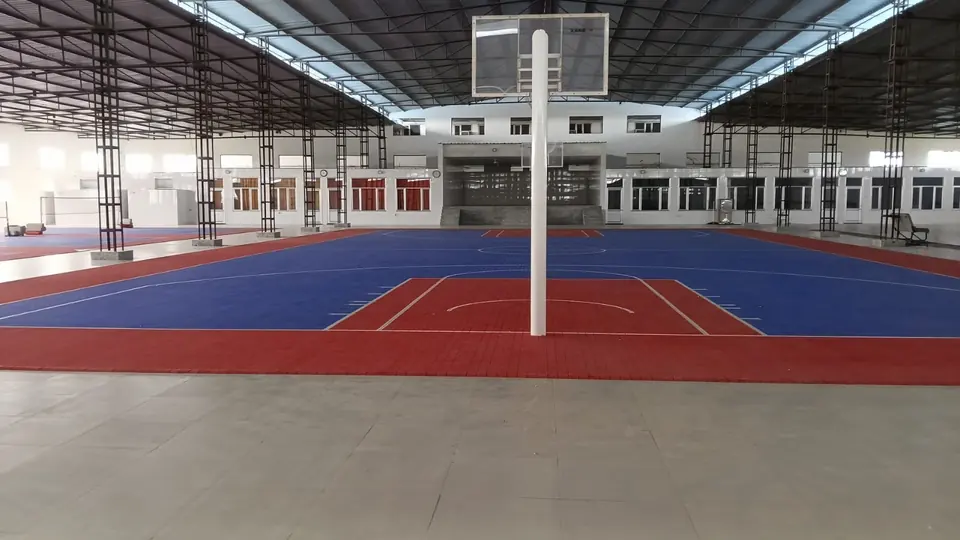 Basketball court on blue and red PP interlocking tiles in a large steel-roofed sports facility, with a basketball pole in the foreground and more courts alongside.