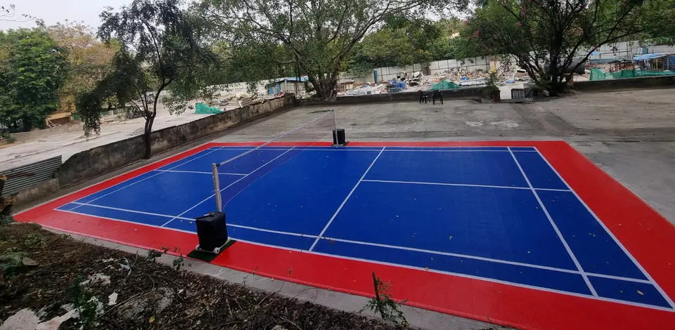Outdoor badminton court on blue PP interlocking tiles with a red surround and white lines, with portable net posts, laid on an open concrete area among trees.