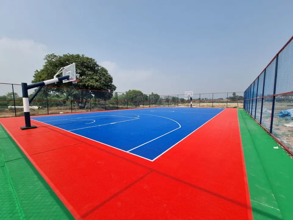 Full outdoor basketball court on blue and red PP interlocking tiles with a green outer edge, two basketball poles and chain-link fencing under a clear sky.