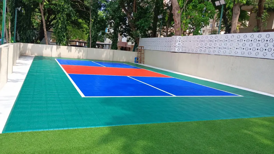 Outdoor pickleball court on blue, orange-red and green PP interlocking tiles with white lines, set between walls and artificial turf among trees.