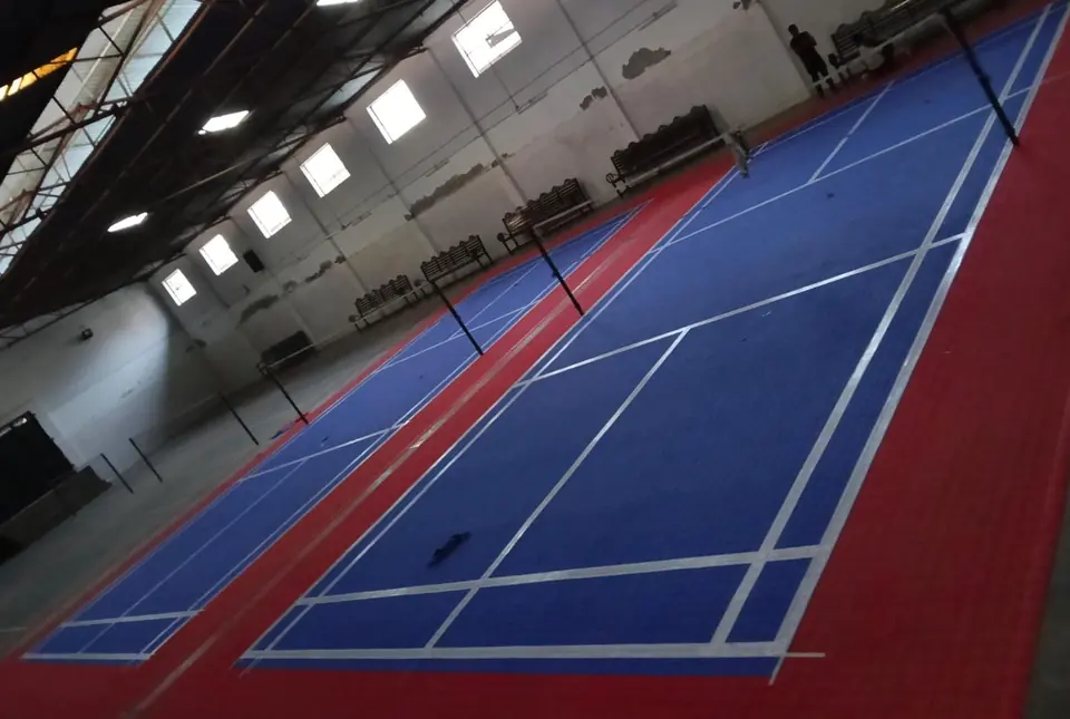 Two indoor badminton courts on blue PP interlocking tiles with red surrounds and white lines, in a hall with high windows and benches.