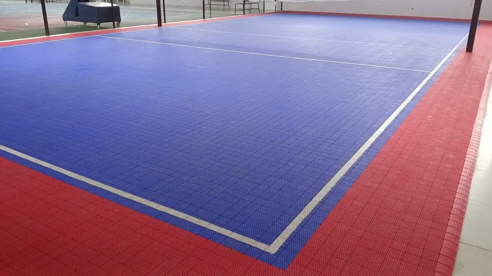 Volleyball court on blue PP interlocking tiles with a red surround and white lines, with tall net posts, inside a covered sports facility.