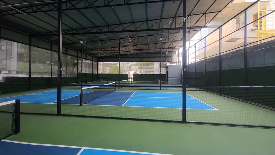 Wide view of the indoor pickleball courts at Rayadurgam, Hyderabad: blue courts with a green surround and nets, divided by netting under a steel-truss roof.