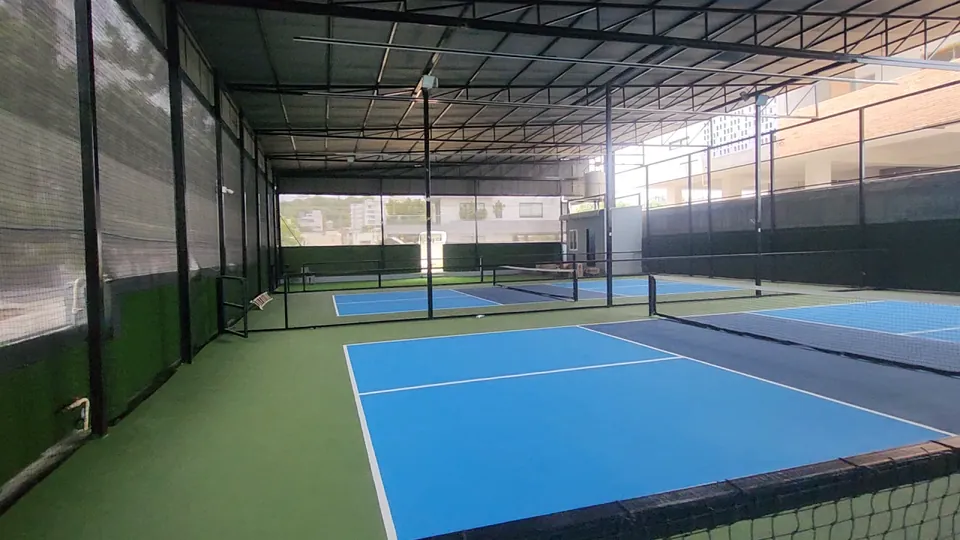 Indoor pickleball court at Rayadurgam with light and dark blue playing areas, white lines and a green surround, with mesh screens along the sides of the shed.