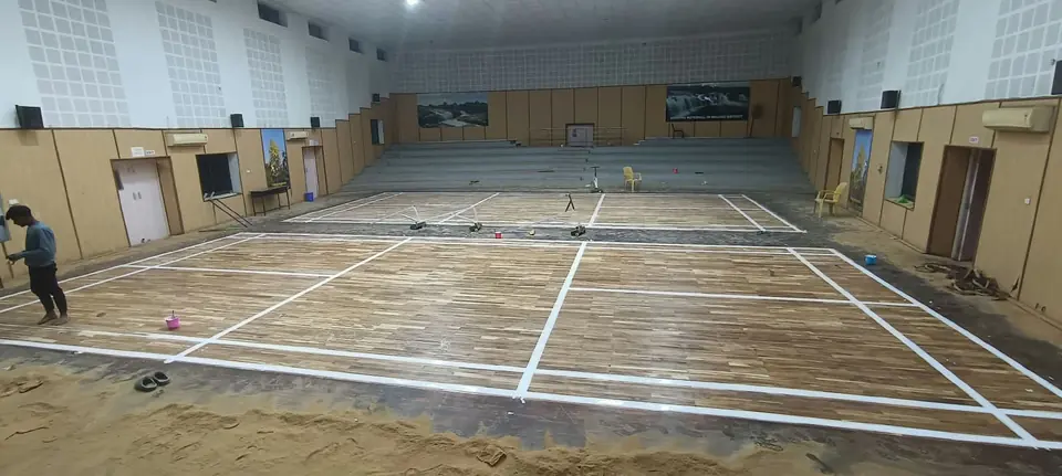 Teak wooden badminton courts being marked out with white line tape in an auditorium hall with tiered seating.