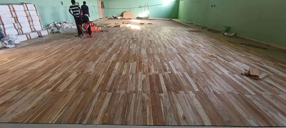 Kelysports team laying teak floor boards across an indoor hall with mint-green walls, with tools and materials nearby.