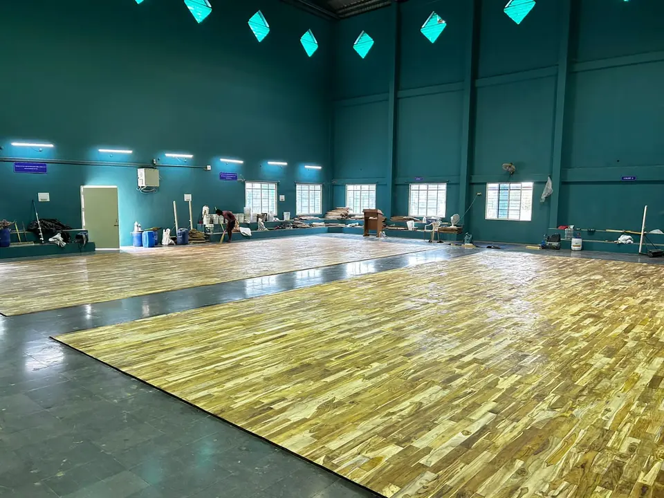 Two teak wooden badminton court floors laid side by side on a stone base in a tall teal-walled sports hall.