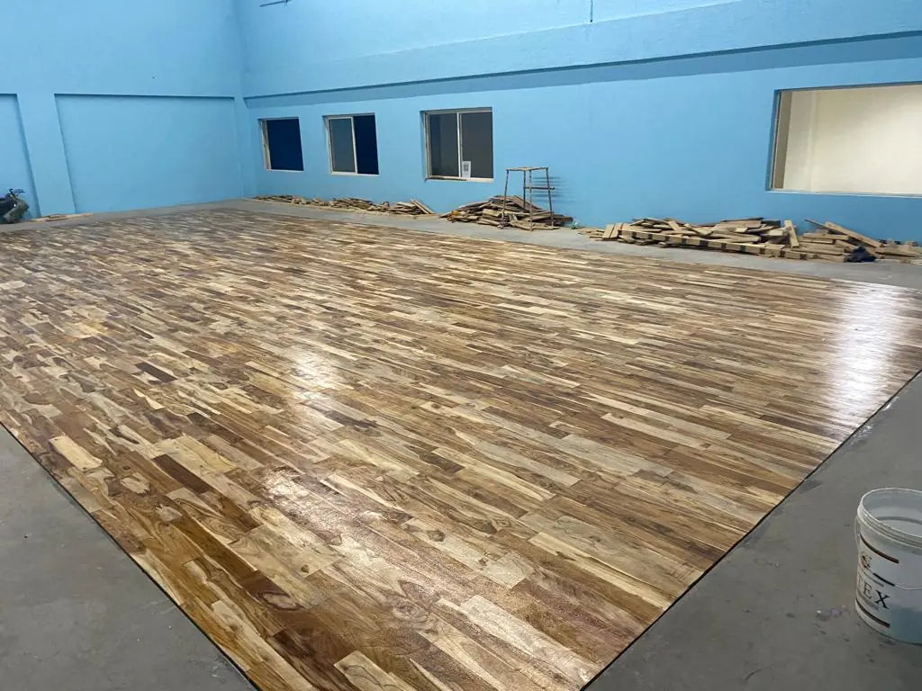 Freshly finished teak wooden floor in a blue-walled indoor hall, with leftover timber offcuts stacked along the wall.
