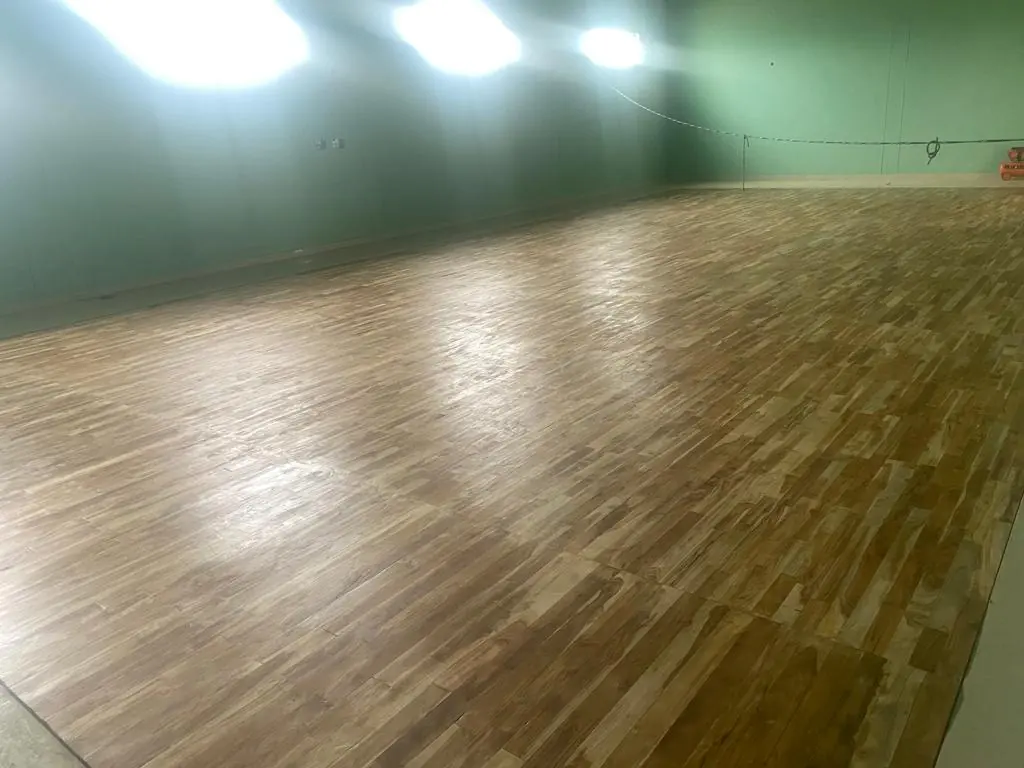 Polished teak wooden sports floor reflecting bright overhead lights in an indoor hall with green walls.